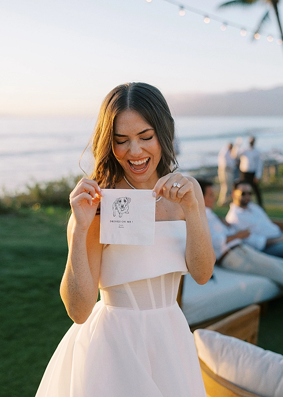 Bride portrait holding a paper card, laughing in a strapless wedding dress with engagement ring, by an ocean lawn with string lights