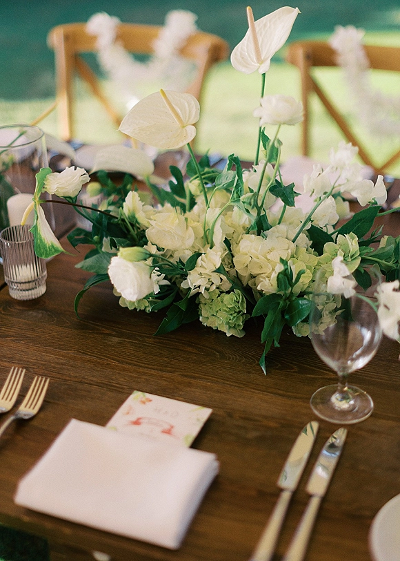 Reception centerpiece with a white floral centerpiece of anthuriums and hydrangeas on a wood dining table with place cards and clear goblets outdoors