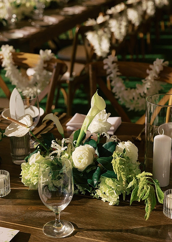 Reception tablescape with a wedding table centerpiece of white hydrangeas, roses, and greenery garland on a sunlit lawn with chairs behind