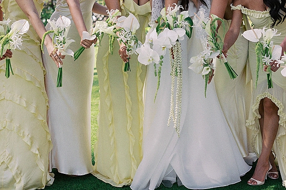 Bridesmaid bouquets with white orchid blooms and calla lilies, trailing stems and ribbons held by women in yellow dresses on a garden lawn