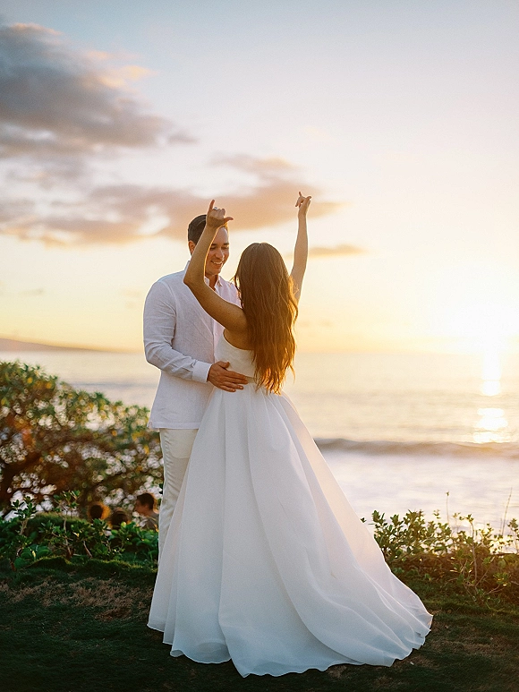 Couple portrait in a sunset wedding photo, bride twirling flowy dress and showing wedding ring as groom embraces her by the ocean shore
