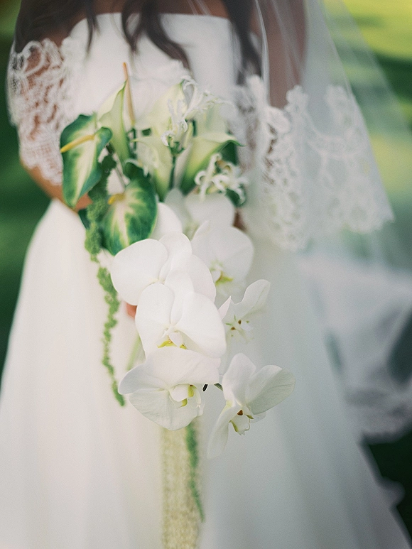 Bridal bouquet of white orchids with green calla lilies cascading over a lace veil, held against a strapless dress in outdoor greenery