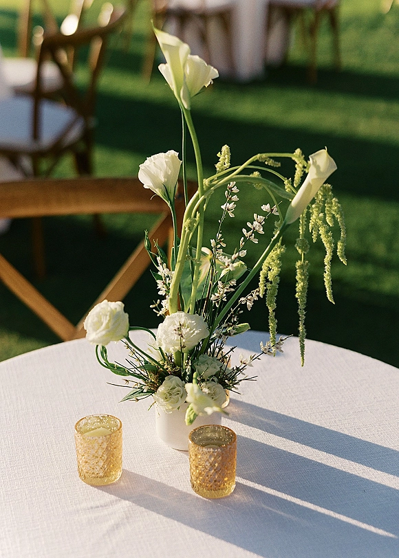 Wedding centerpiece with tall floral centerpiece of calla lilies and white blooms in a white vase, with amber votives on an outdoor lawn table