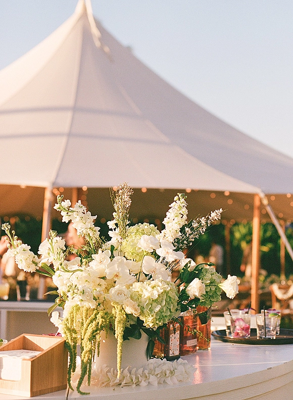 Wedding centerpiece with white floral centerpiece in a white vase on a bar counter, framed by cocktail glasses and amber votives under string lights