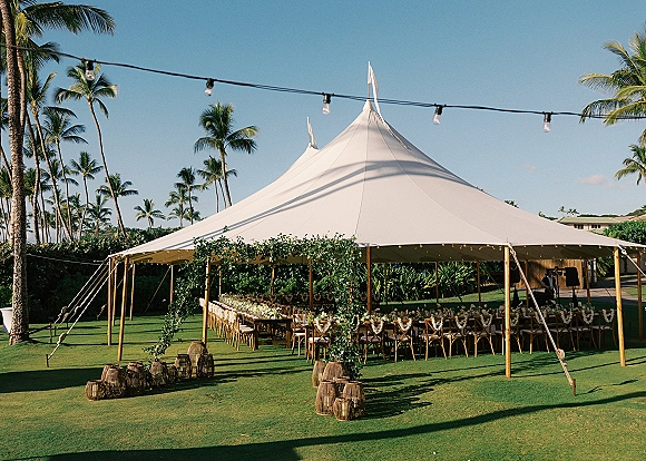 Wedding reception tent sailcloth tent reception with string lights and greenery garlands over long banquet tables on a lawn with palm trees