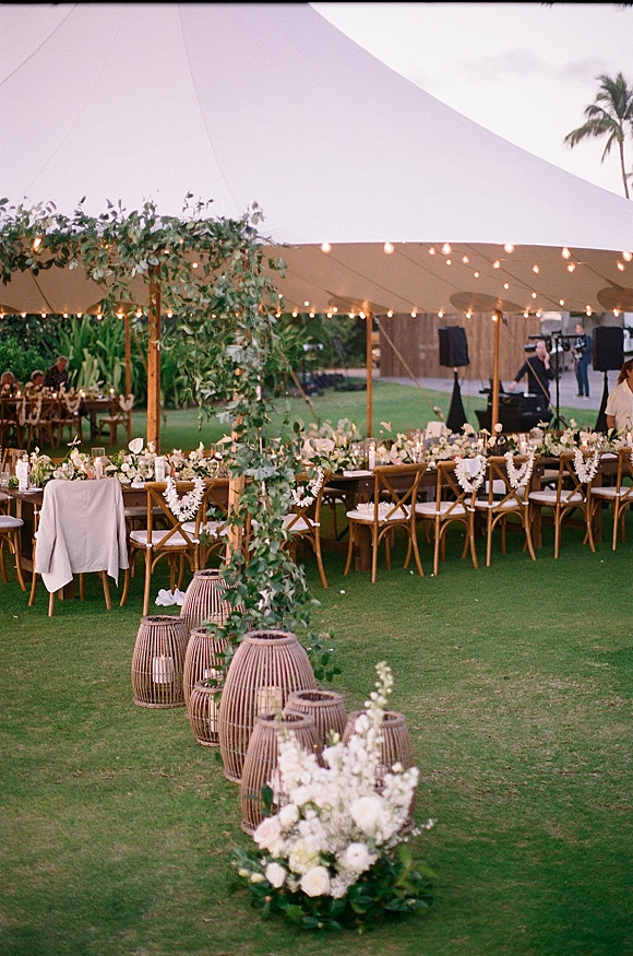 Tent wedding reception under a sailcloth tent with string lights, long tables, cross back chairs, and candlelit white floral garlands on a lawn