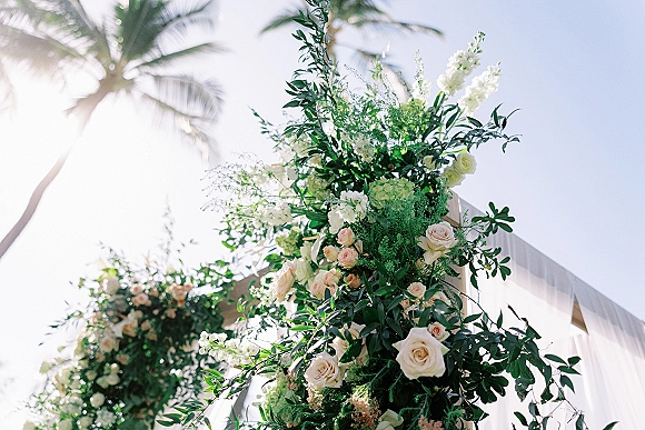 Ceremony arch flowers with white and blush roses and greenery, draped in white fabric, set against sunlit palm trees and sky
