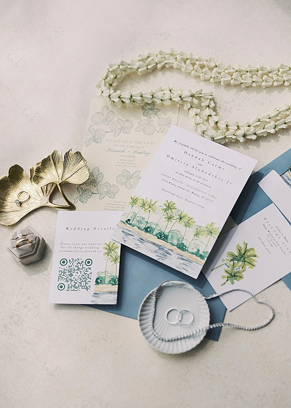 Invitation suite wedding invitation flat lay with watercolor cards, blue envelopes, rings, pearl necklace, and floral lei on a neutral tabletop