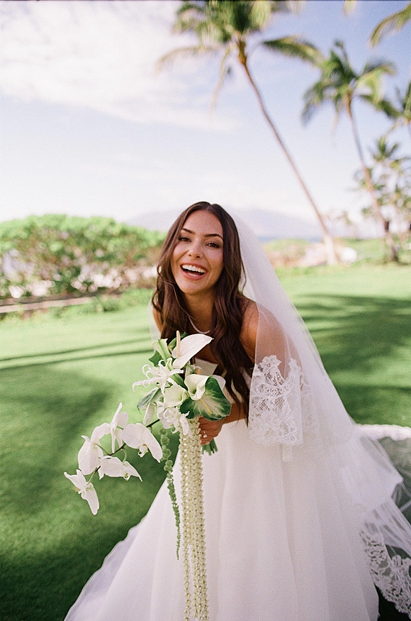 Bridal portrait of a smiling bride holding bouquet of cascading white orchids and calla lilies, in lace veil on a palm-lined lawn under blue sky