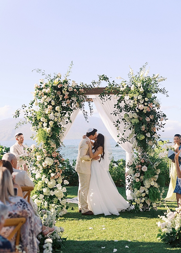 Ceremony kiss moment at an outdoor wedding ceremony, bride and groom under a white draped floral arch with ocean and mountains behind