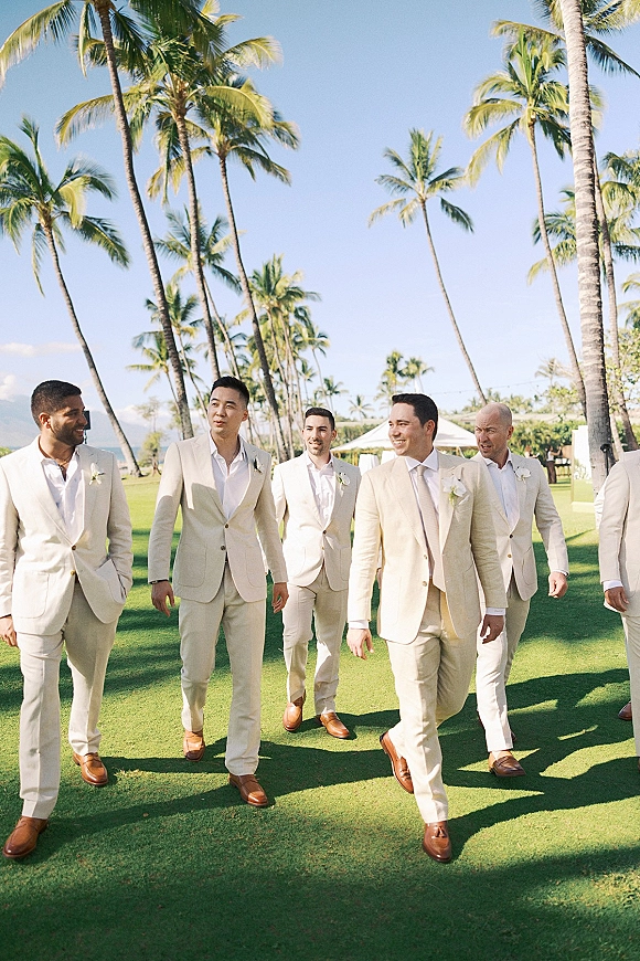Groomsmen portrait of five men in beige suits with boutonnières and brown shoes, laughing on a lawn with palm trees and mountains behind