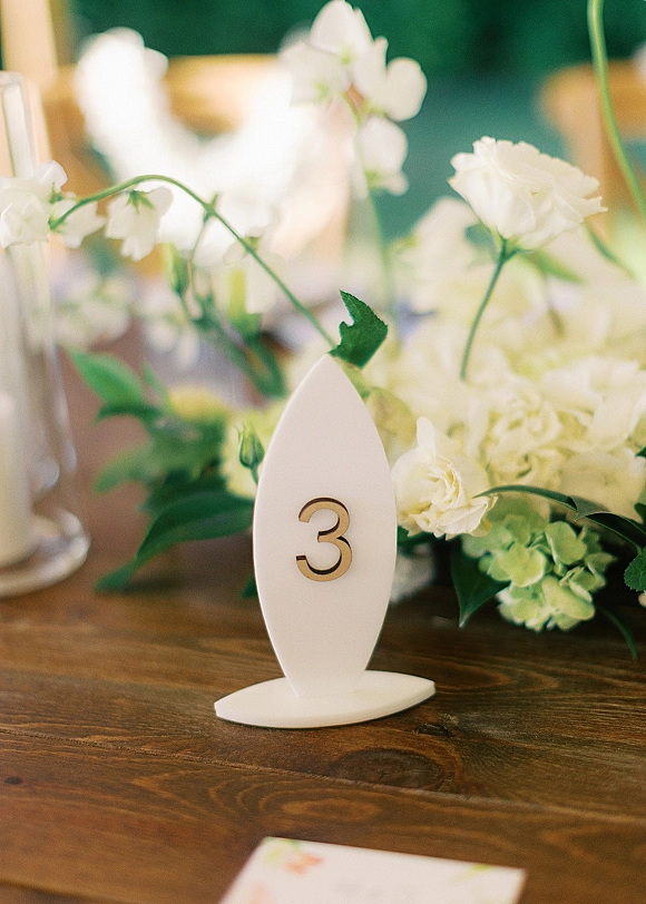 Wedding table number sign beside a white floral centerpiece with greenery in a vase on a wood table, blurred reception space behind