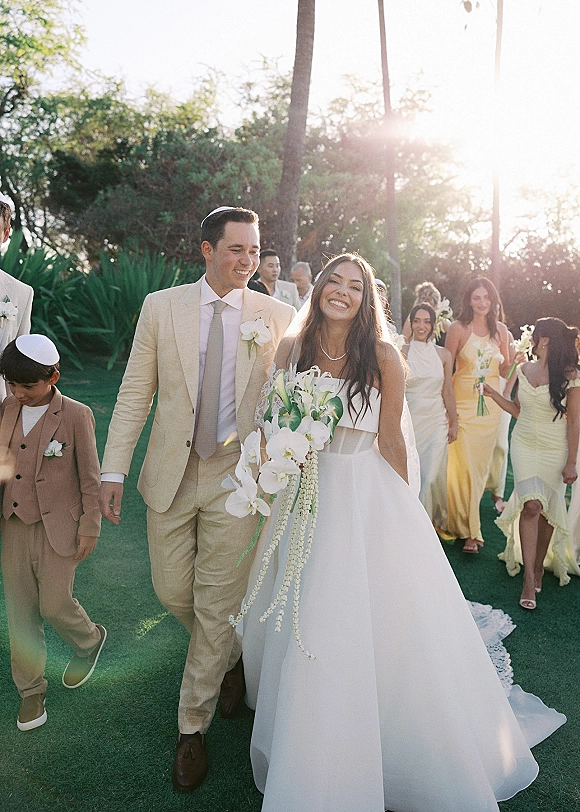 Wedding recessional as bride and groom walking hand in hand, bride holding a cascading orchid bouquet, wedding party behind on a sunny lawn with palm trees