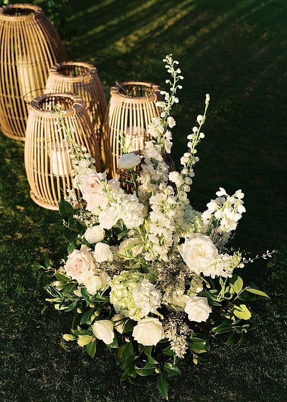 Ceremony aisle florals with ground floral arrangement of white and blush roses, greenery, and rattan lanterns with candles on a grassy lawn