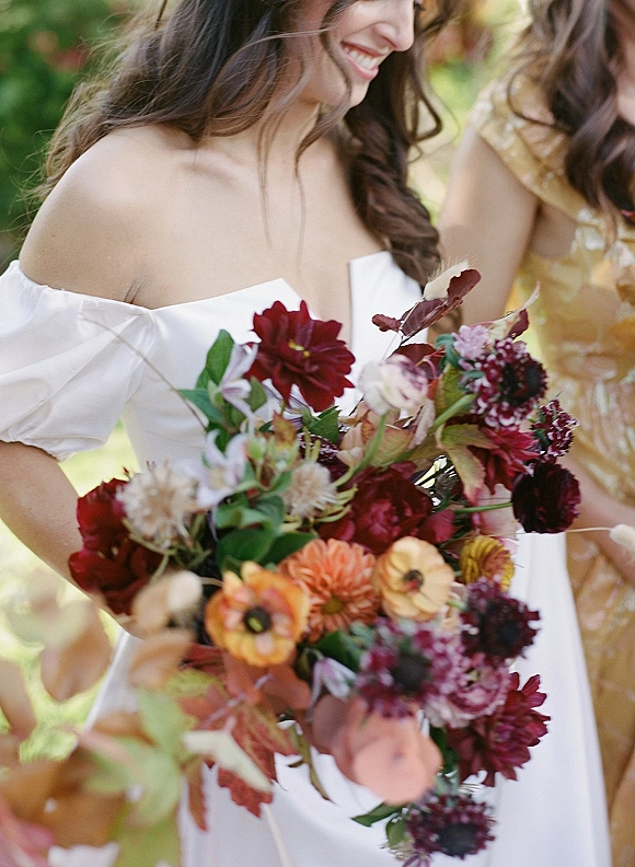 Bridal bouquet with burgundy bridal bouquet blooms in orange and greenery, ribbon-wrapped, held against sunlit outdoor bokeh backdrop