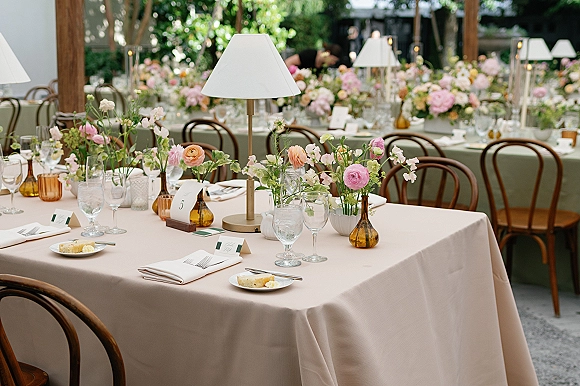 Reception tablescape with pastel floral centerpieces, table lamps and amber bud vases on blush linens at an outdoor garden patio banquet table