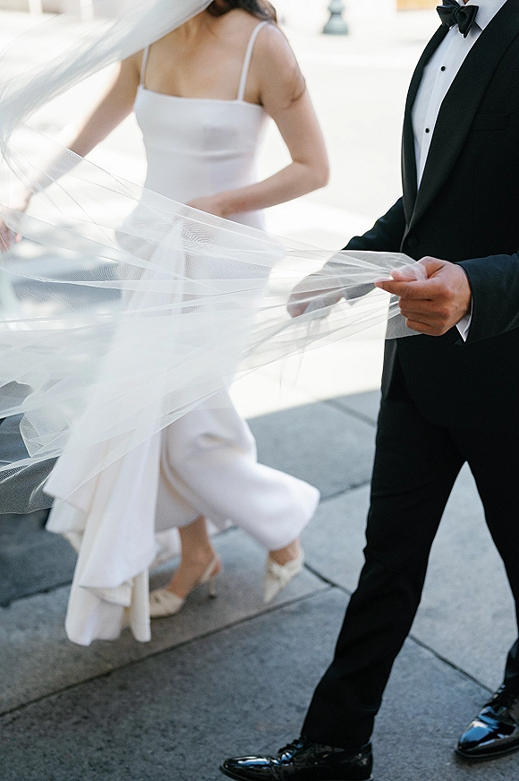 Couple portrait of bride and groom walking with a wedding veil blowing between them, bride in short gown and heels on a paved walkway