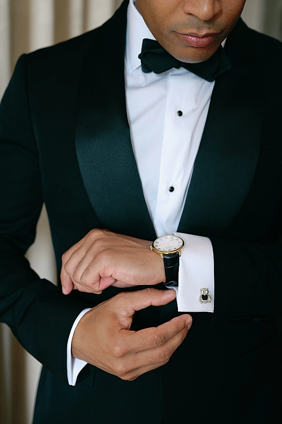 Groom getting ready in a black tuxedo, adjusting cufflinks and wristwatch over a white dress shirt beside neutral curtains
