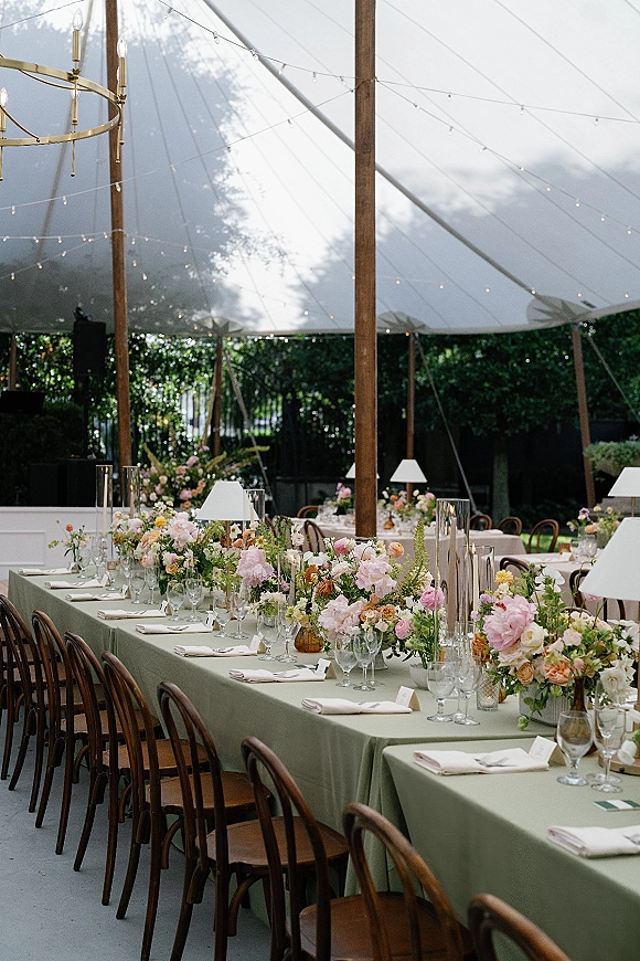 Reception tablescape with long banquet table in a sailcloth tent, sage green linens, floral centerpieces, taper candles, and chandeliers overhead