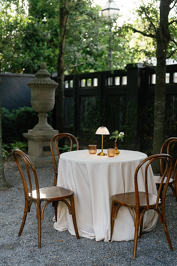 Cocktail table setup with white tablecloth, bistro chairs, cordless lamp, bud vase florals, and votive candles on a gravel patio by garden trees
