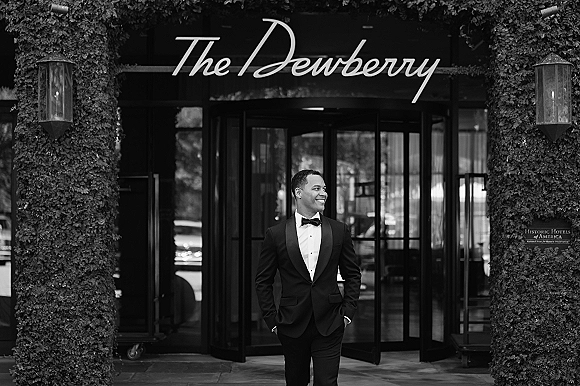 Groom portrait in a black tuxedo and bow tie, hands in pockets, smiling outside a hotel entrance by a revolving door and ivy wall