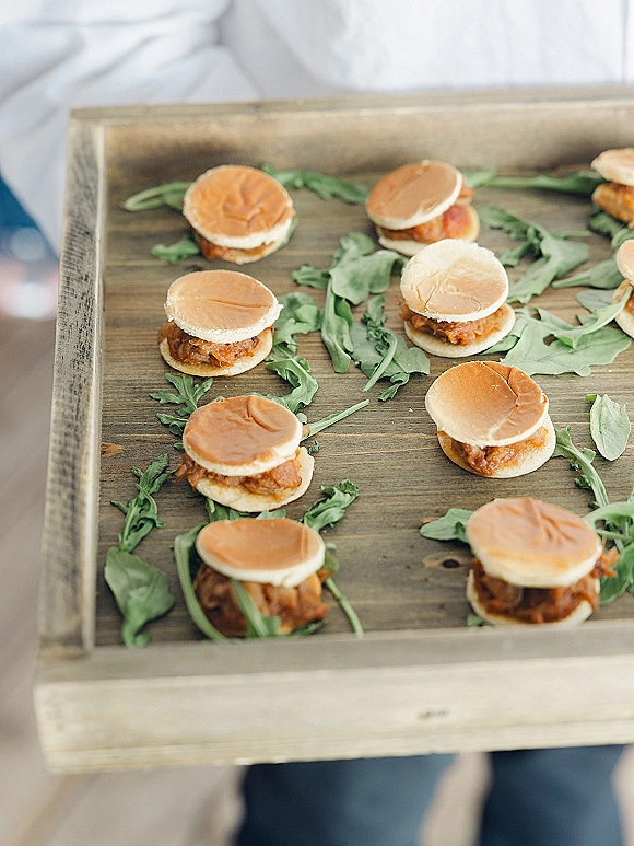 Wedding appetizer sliders on a wooden serving tray with arugula greens, held by a server in a white shirt in a blurred indoor setting