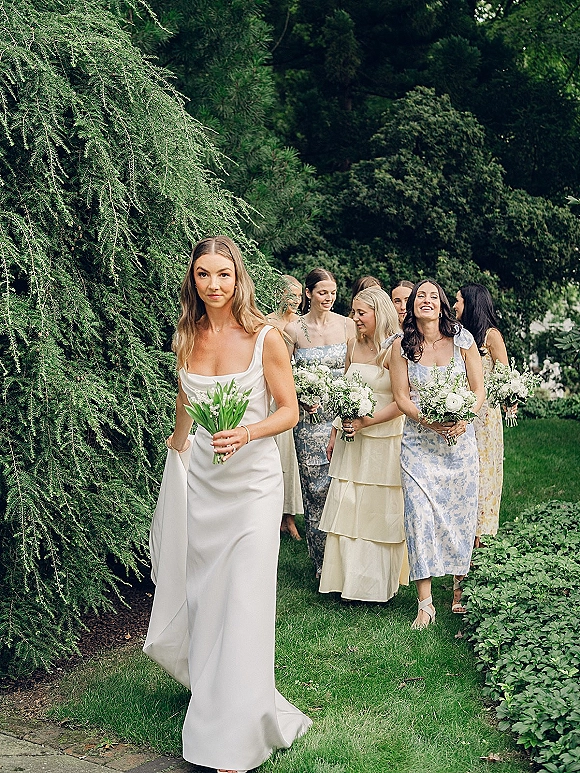 Bridesmaid procession with bridesmaids walking in mixed dresses, holding white calla lily bouquets on a garden lawn amid lush greenery