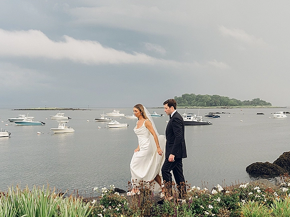 Couple portrait of bride and groom walking along a rocky shoreline, veil in the wind beside ocean boats under a cloudy sky
