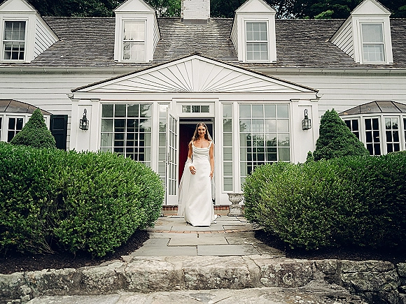 Bridal portrait of a bride walking outside in a sleek satin wedding dress and long veil by a white house with French doors and hedges