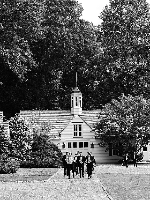 Groomsmen portrait with black tie groomsmen in tuxedos and bow ties walking on a driveway beside a white chapel with cupola and trees