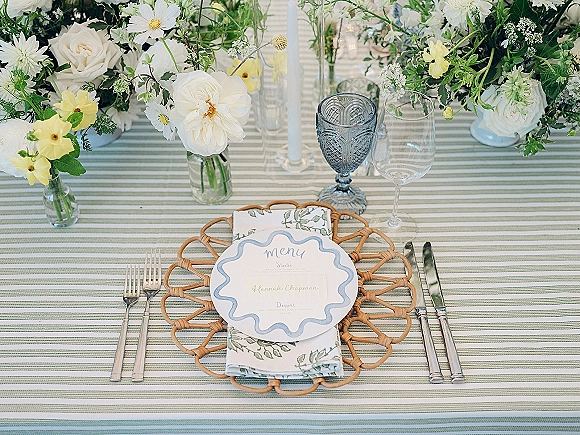 Wedding tablescape reception place setting with striped tablecloth, rattan placemat, blue goblet, taper candle, and white-yellow florals