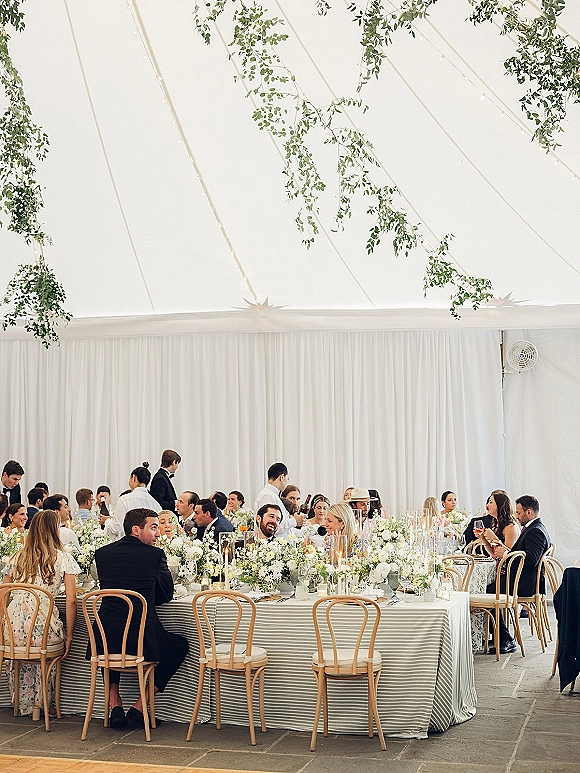 Reception tablescape with long banquet table wedding styling, striped tablecloth, white florals, greenery garlands, and taper candles under string lights