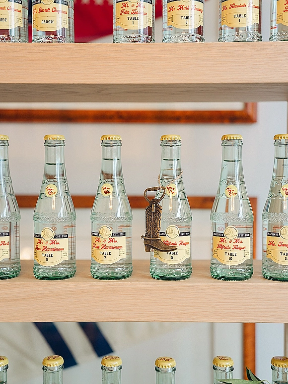 Wedding escort cards on glass soda bottles with custom labels, twine and a mini boot charm, arranged on wooden shelves against a white wall