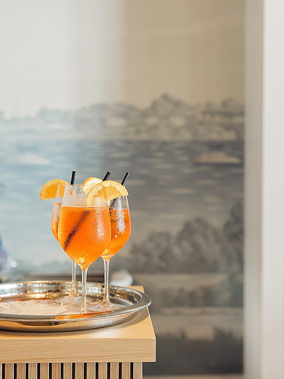 Wedding cocktails on a metal tray, orange slice garnish and black straws in stemmed glasses on a wood bar cart before a mural wall art backdrop