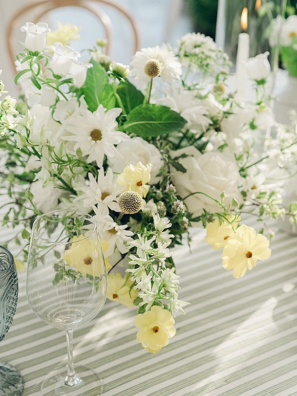 Wedding centerpiece with white and yellow flowers and greenery beside a votive candle and wine glass on a striped tablecloth outdoors
