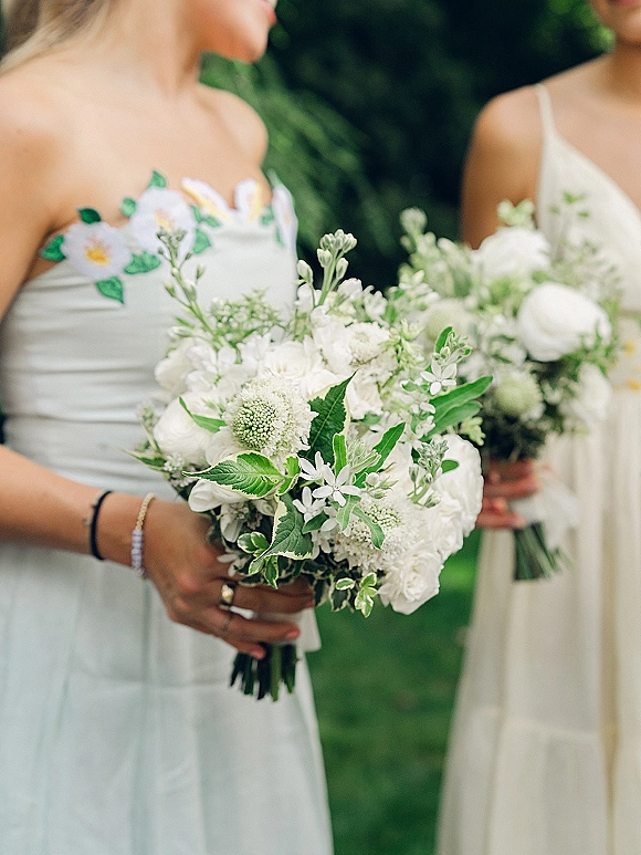 Bridesmaid bouquets of white and green flowers with ribbon wraps held by bridesmaids in light blue dresses on a garden lawn backdrop