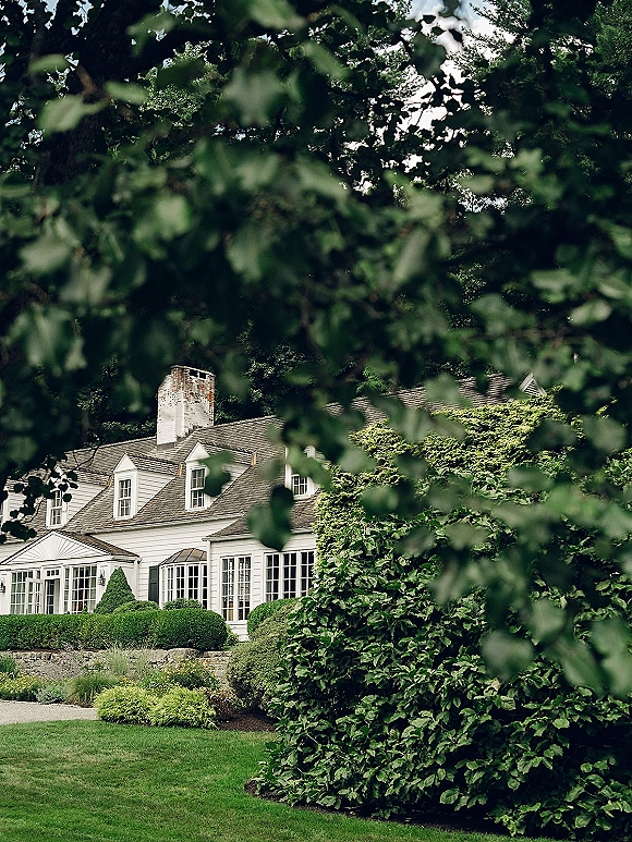 Wedding venue exterior with ivy accents on a white house, stone wall and chimney, framed by trees and manicured lawn landscaping