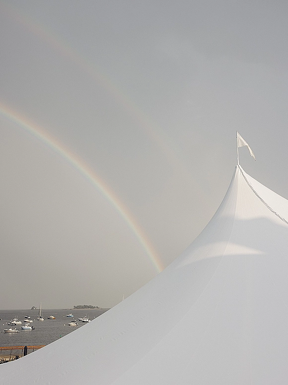 Wedding tent with a sailcloth wedding tent flag, set on the waterfront with boats in the harbor and a rainbow under cloudy skies