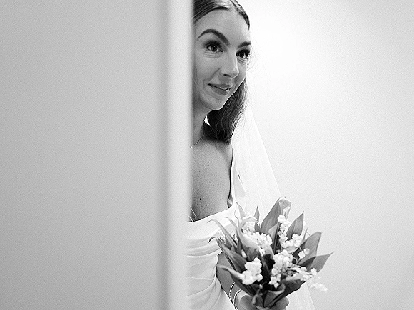 Bridal portrait in black and white of a bride peeking through a curtain, holding a lily of the valley bouquet, veil over shoulder