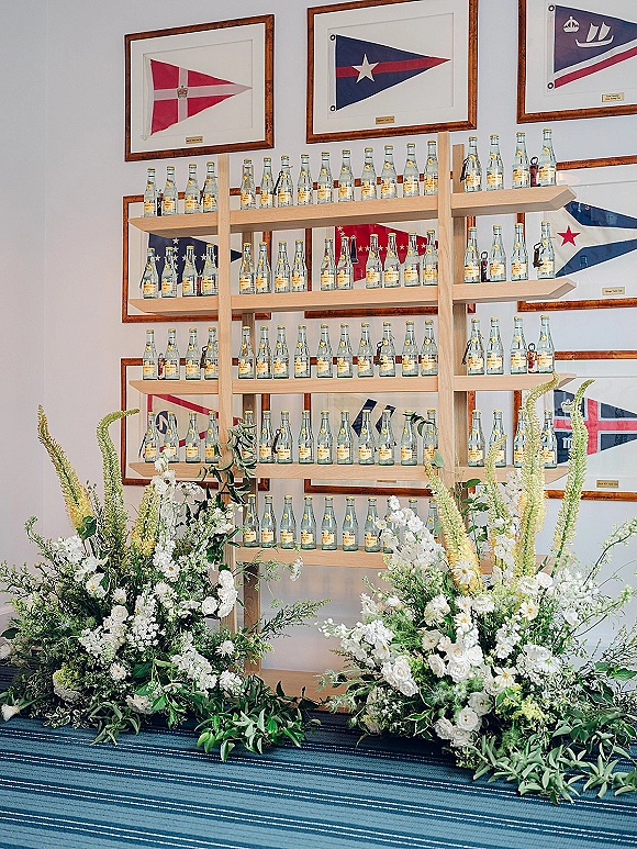 Wedding escort card display with bottle escort cards on wood shelving, paired with greenery and florals beneath framed nautical flags