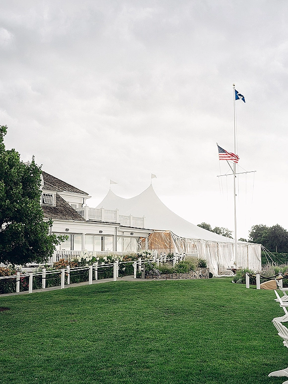 Wedding reception tent with sailcloth canopy and string lights, clear sidewalls on a green lawn near a white clubhouse under cloudy skies