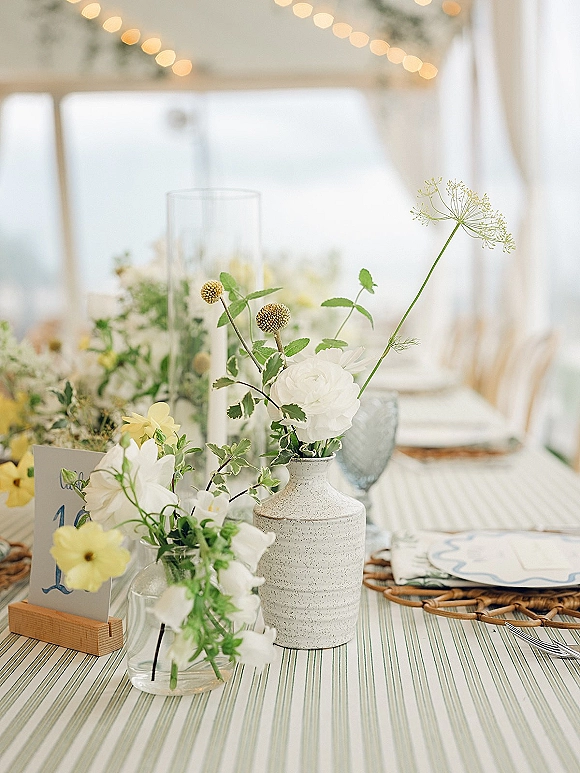Reception tablescape with wedding table centerpiece in mixed bud vases, wildflowers and greenery on striped linens under tent string lights