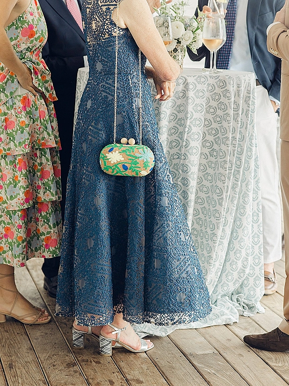 Wedding guest outfit featuring a blue lace wedding guest dress with floral clutch and patterned block heels beside a cocktail table at indoor reception