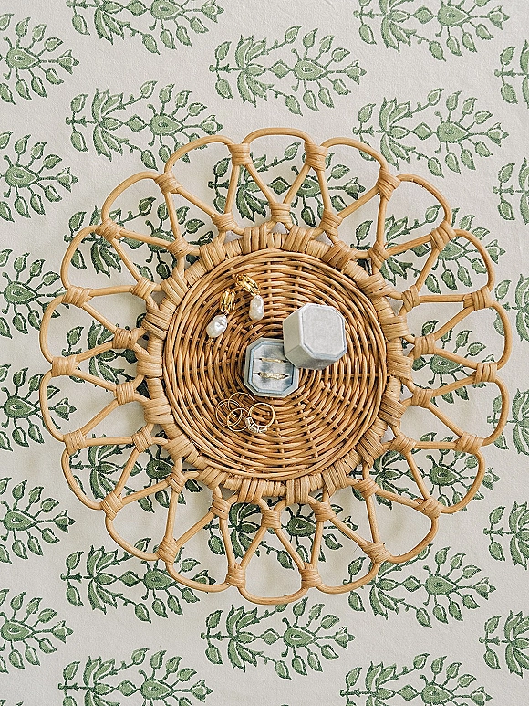 Wedding rings and engagement ring close up in a ring box beside pearl drop earrings on a rattan tray over green floral fabric