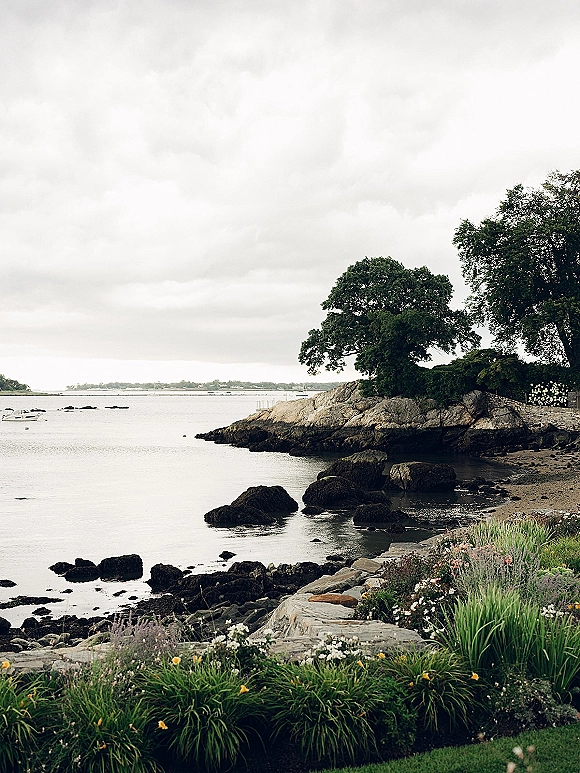 Coastal wedding venue with a seaside flower garden accent beside a stone wall, overlooking rocky shoreline and ocean under cloudy sky