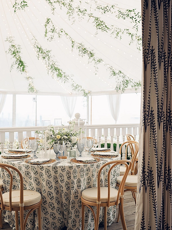 Reception tablescape with a round wedding reception table in a white tent, featuring woven chargers, glass goblets, greenery, string lights.