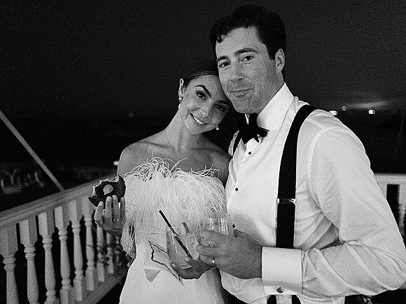 Wedding couple portrait in a black and white wedding photo, bride in strapless feather-trim dress holding doughnut and drinks on a deck at night