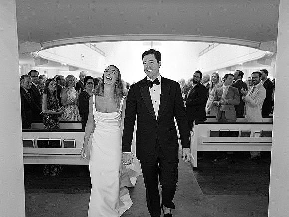 Wedding recessional with newlyweds walking down aisle holding hands, bride in long veil and groom in tuxedo as guests clap in church pews