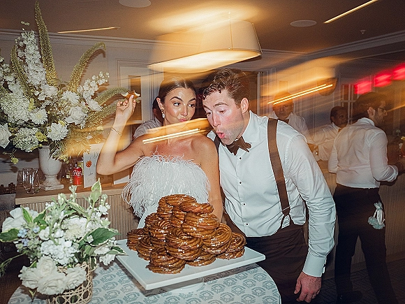 Wedding cake moment as bride in a feather dress and groom in bow tie and suspenders blow on a stacked pastry cake under a chandelier