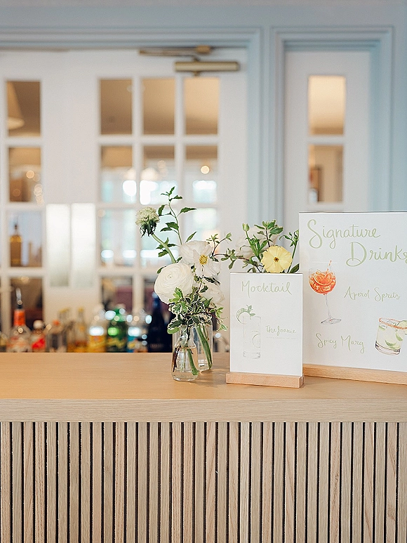 Signature drinks sign and wedding signature drink menu displayed on a bar counter with a white floral vase and greenery in an indoor bar area